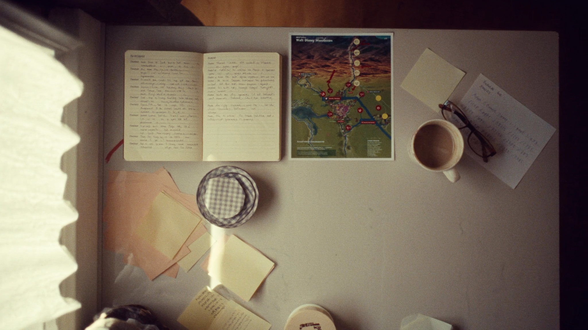 Top-down view of a kitchen table. Open notebook with handwritten travel notes, a printed Walt Disney World park map with hand-drawn arrows and dot stickers, a half-drunk cup of milky tea, sticky notes in muted earth tones, reading glasses.