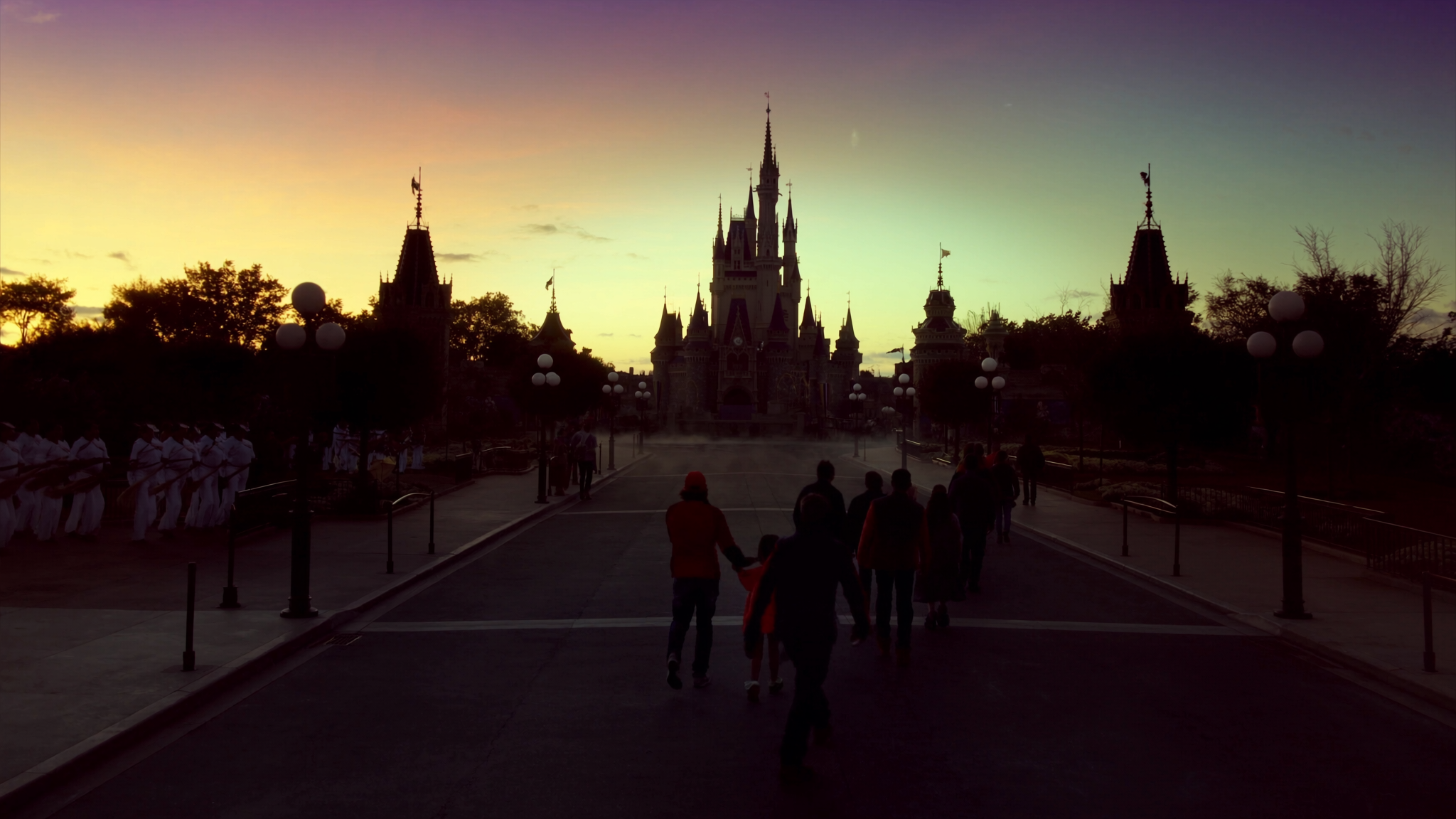 Rope drop sunrise. Family silhouettes walk up an empty Main Street USA toward Cinderella Castle, sky transitioning from deep aubergine to teal to apricot at the horizon.
